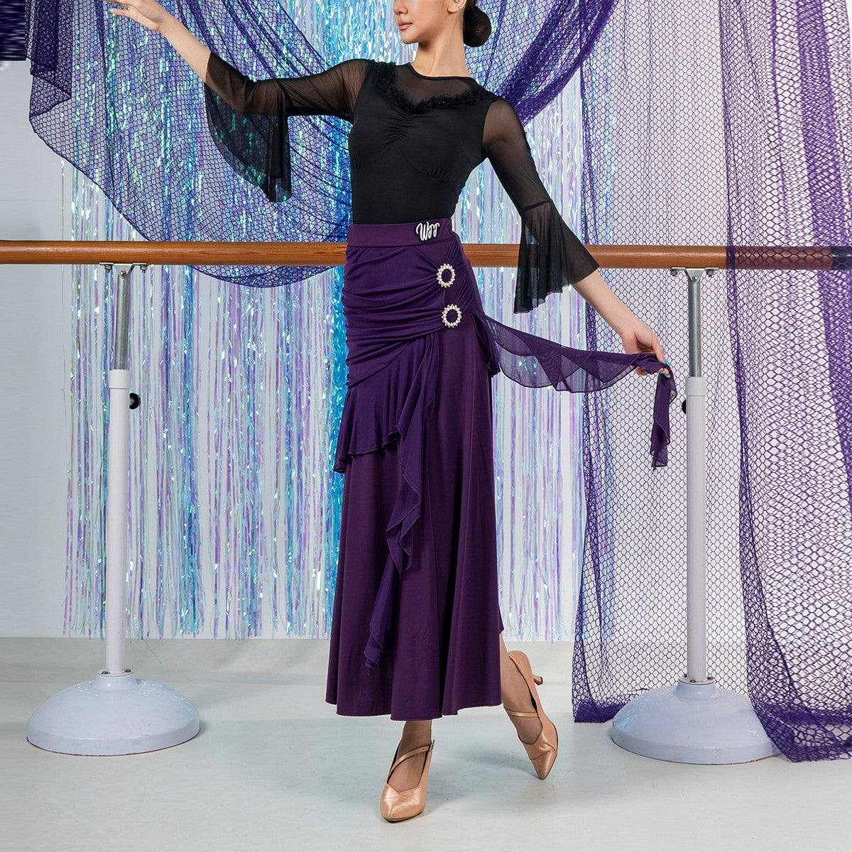 Purple ballroom dance skirt with ruched panel and rhinestone buckles, front view at the barre