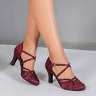 Woman wearing burgundy satin closed toe ballroom dance shoes with black rhinestone straps and rhinestone heels